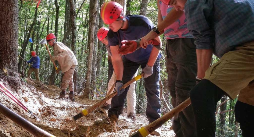 A group of people wearing hard hats use tools to work on a trail as part of a service project